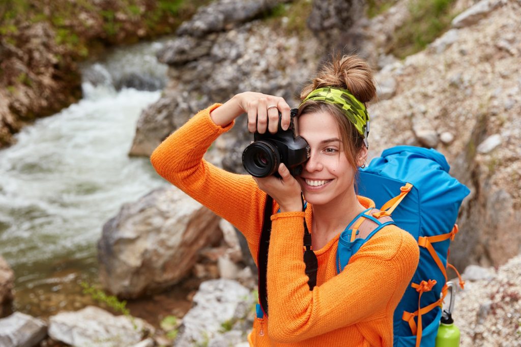 Young female adventurer poses against small river in ravine, holds camera, takes picture of landscap